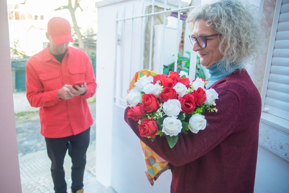 A woman with curly gray hair, wearing glasses, a blue turtleneck, and a maroon cardigan, is holding a bouquet of fresh flowers consisting of red and white roses, wrapped in colorful paper. She is stan
