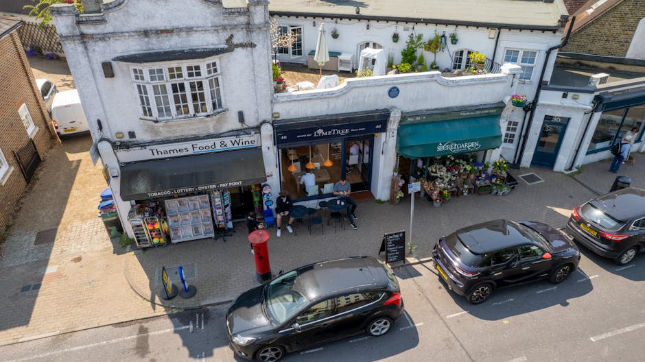 An aerial view of a busy storefront on a street near Thames Ditton station, featuring a flower shop with a green awning labeled 'Secret Garden' and a display of colorful potted plants and floral arran
