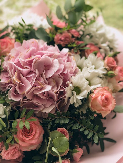 A floral arrangement featuring a large, soft pink hydrangea as the central focus, surrounded by pale peach roses, white daisies with yellow centers, and sprigs of greenery with small, rounded leaves. 
