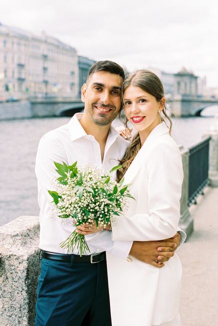 A man and woman stand together outdoors beside the river in Thames Ditton, holding a fresh bouquet arrangement of white baby's breath, lily of the valley, and green foliage. The woman, wearing a white