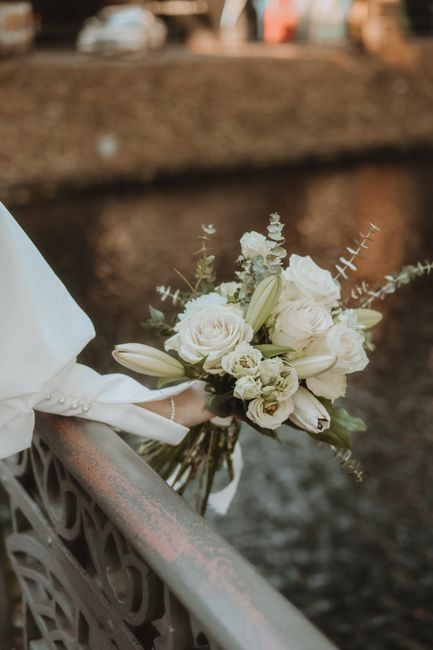 A floral bouquet consisting of white roses, lilies, and other delicate white flowers, accented with light green foliage and silver-grey eucalyptus. The bouquet is held by a person dressed in a white s