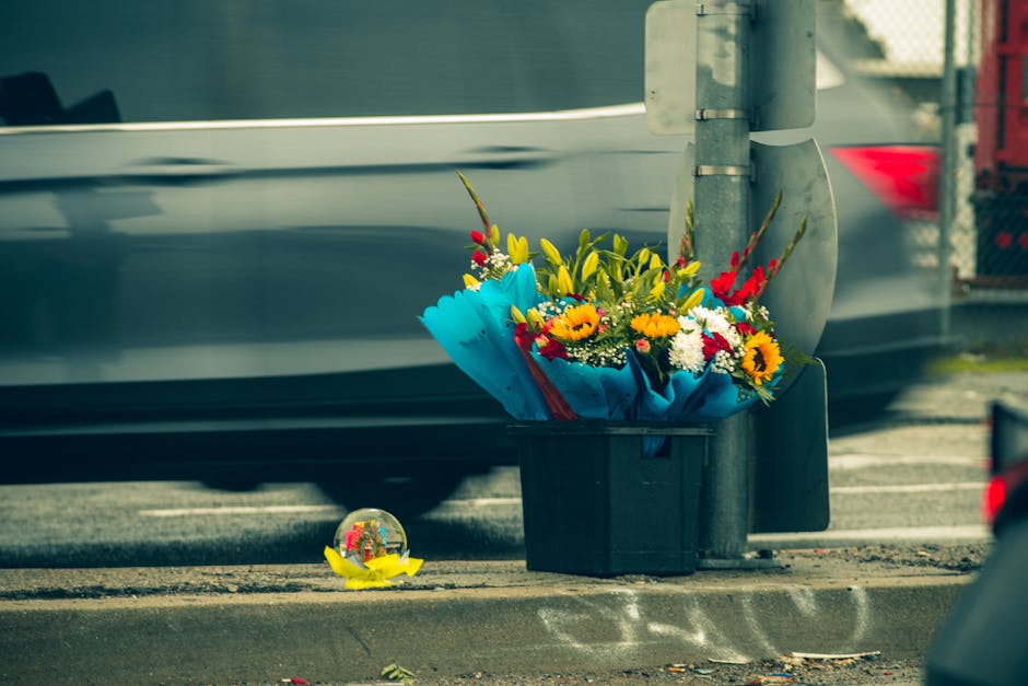 A bouquet of fresh flowers, including sunflowers, red and yellow lilies, and white daisies, wrapped in bright blue paper and placed in a black delivery tub at the side of a road. The arrangement featu